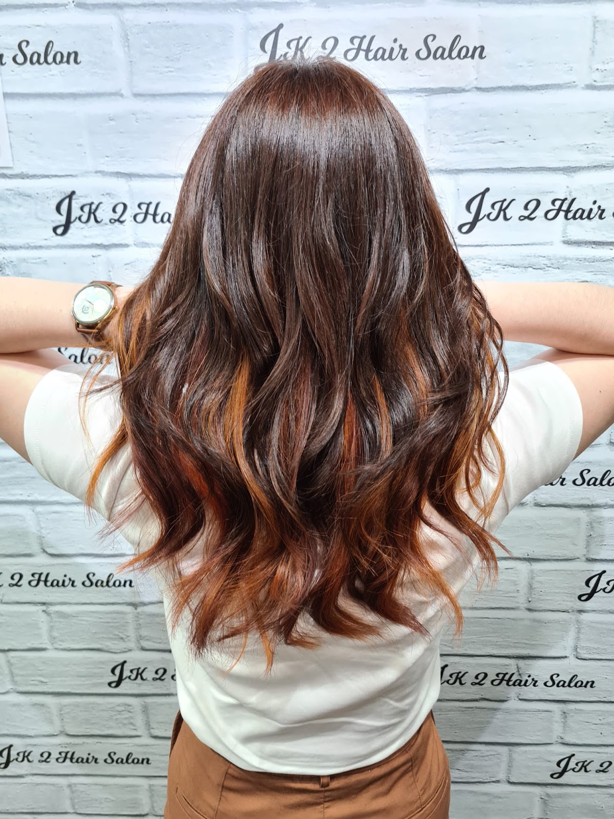 Long waves coloured in a copper and auburn balayage, photographed from behind against the salon's branded wall